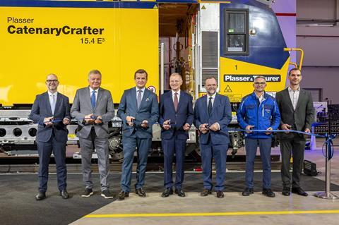 Seven officials and Plasser & Theurer staff cutting a ribbon in front of a yellow CatenaryCrafter track machine at the opening of the Linz factory