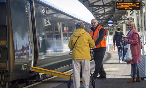Staff member assisting passenger with mobility aid onto train with ramp deployed at a train station